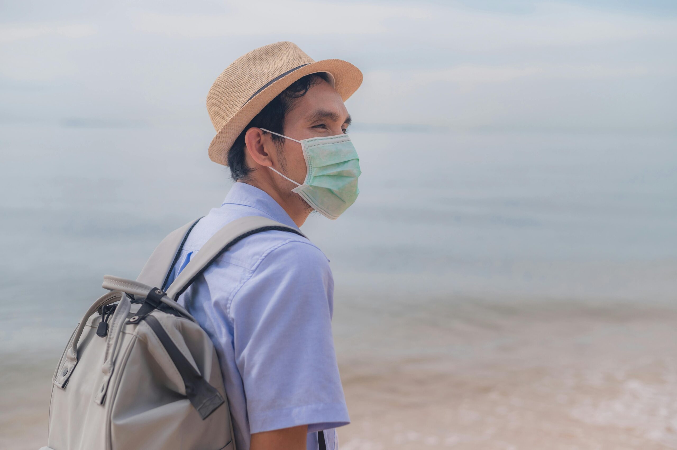 Man wearing a surgical mask and fedora backpacking on a serene beach, embracing the new normal.